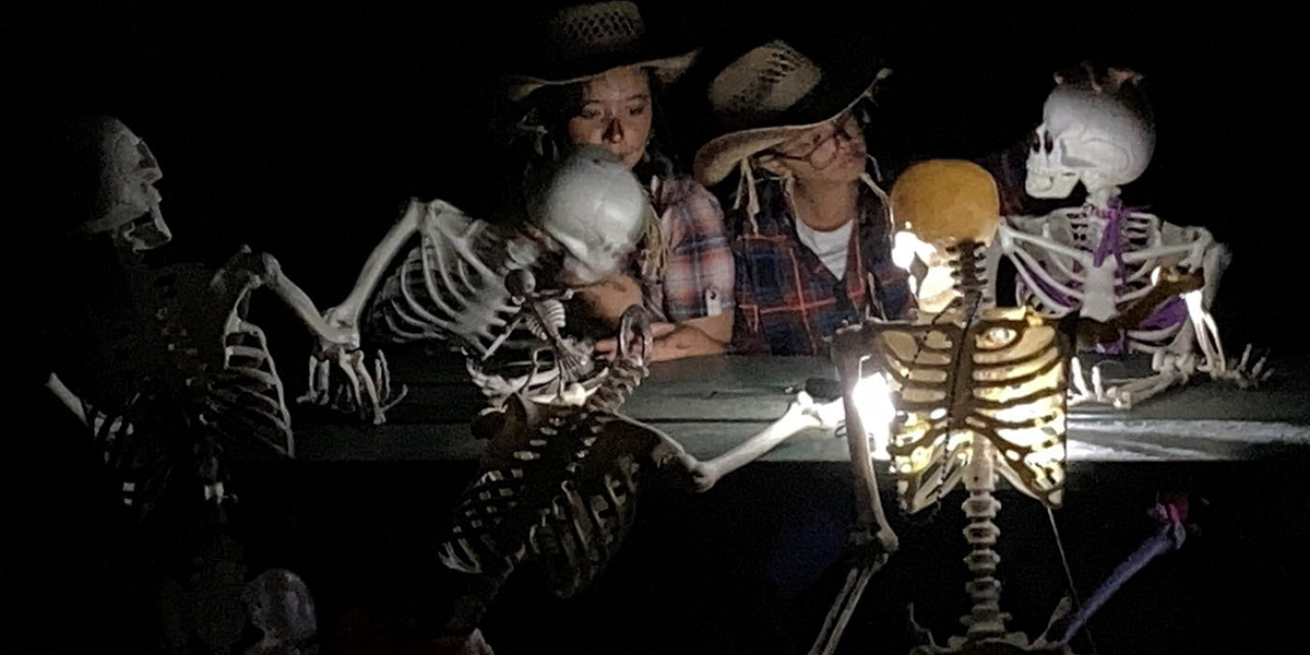 Two girls sit at a picnic table with 3 skeletons